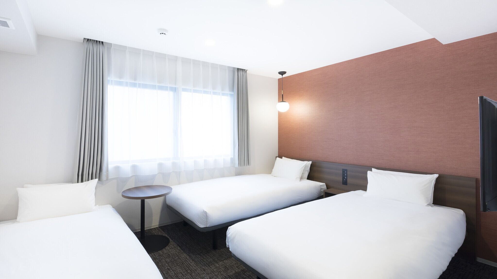 Une chambre d’hôtel avec deux lits simples, une petite table ronde, une grande fenêtre avec des rideaux, un téléviseur fixé au mur et un luminaire au plafond.