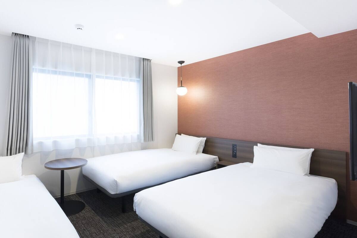 A hotel room with two single beds, a small round table, a large window with curtains, a wall-mounted TV, and a ceiling light.