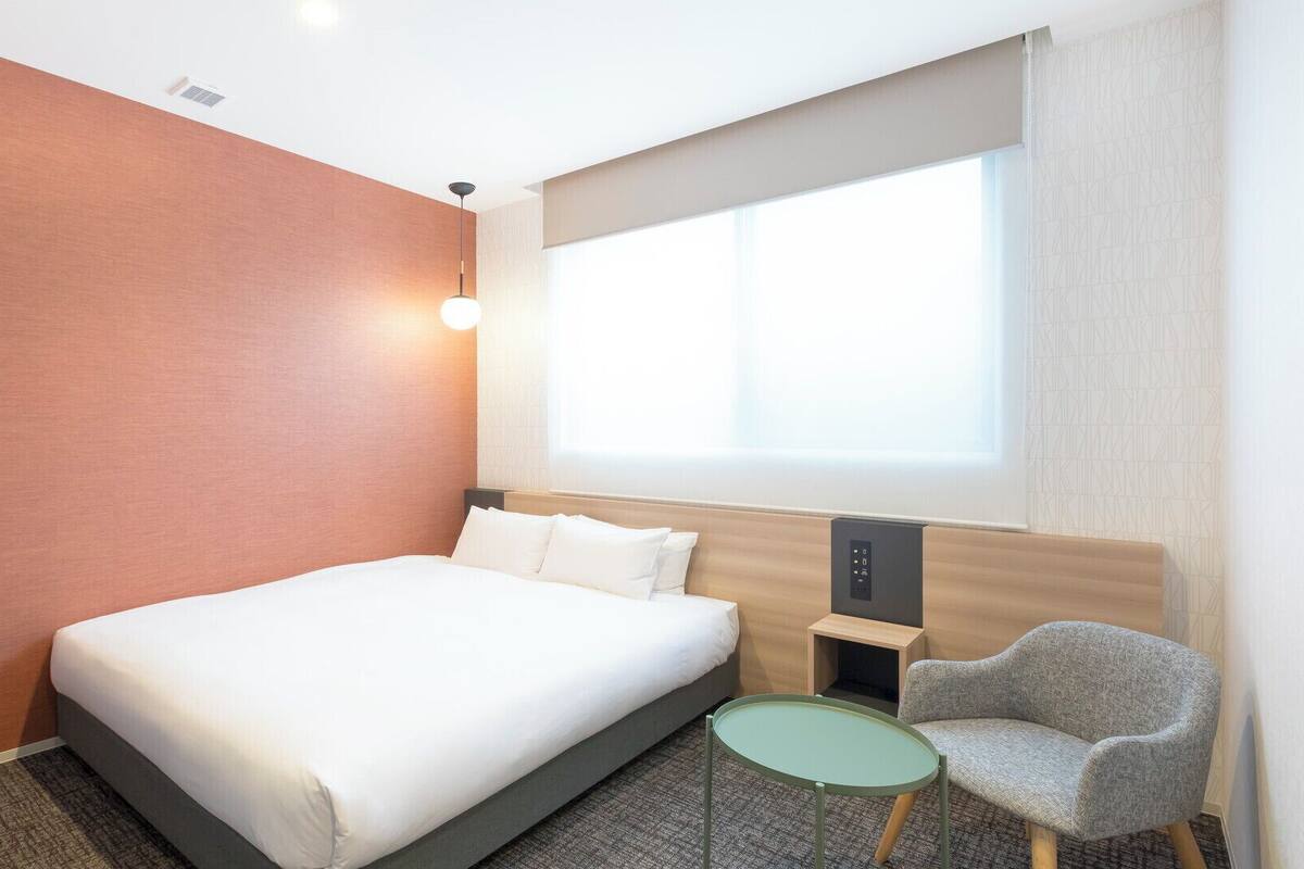 A hotel room with a bed, a small table, a chair, a nightstand, and a window with curtains.