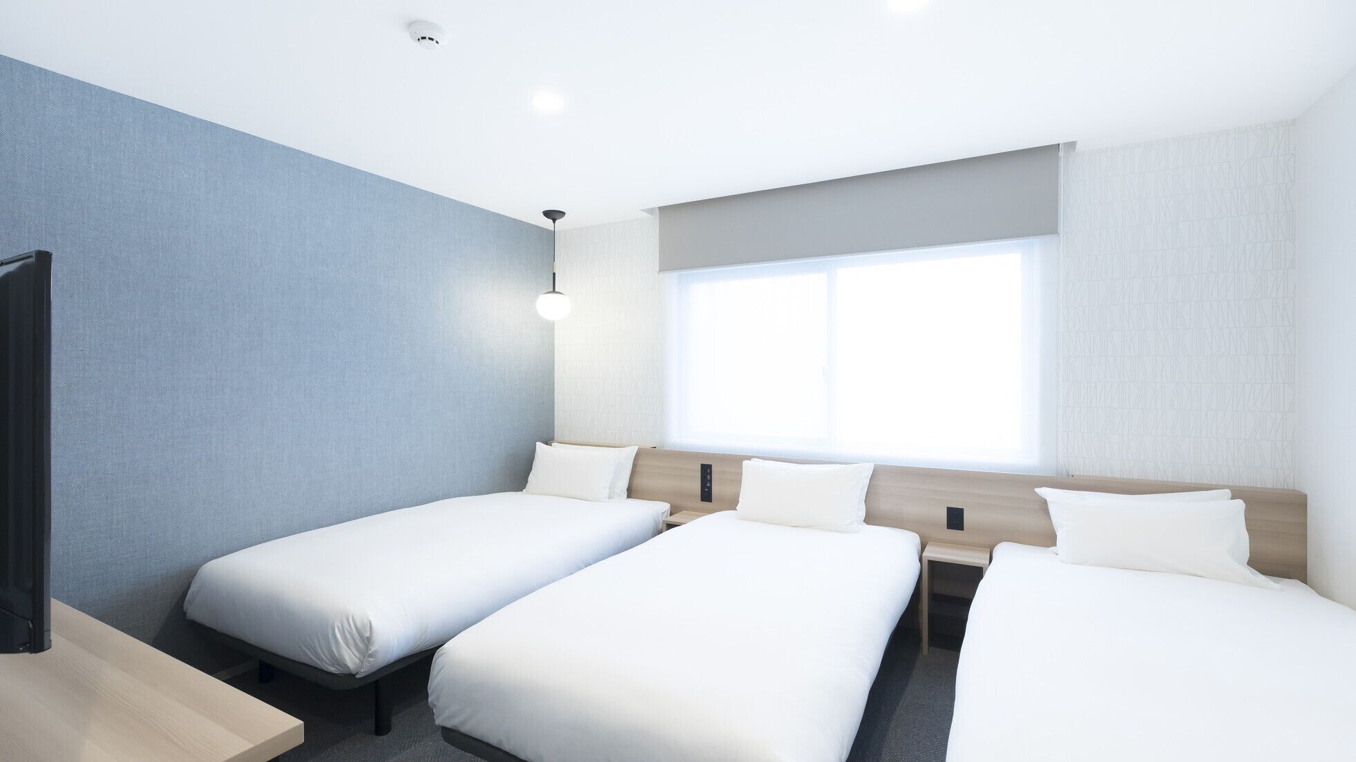 Une chambre d’hôtel avec trois lits simples, un bureau, une chaise, une fenêtre et un luminaire fixé au plafond.