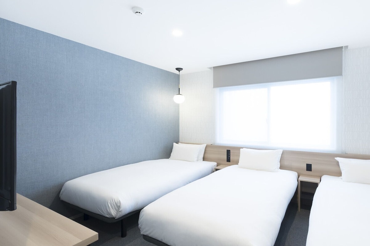 A hotel room with three single beds, a desk, a chair, a window, and a ceiling-mounted light fixture.
