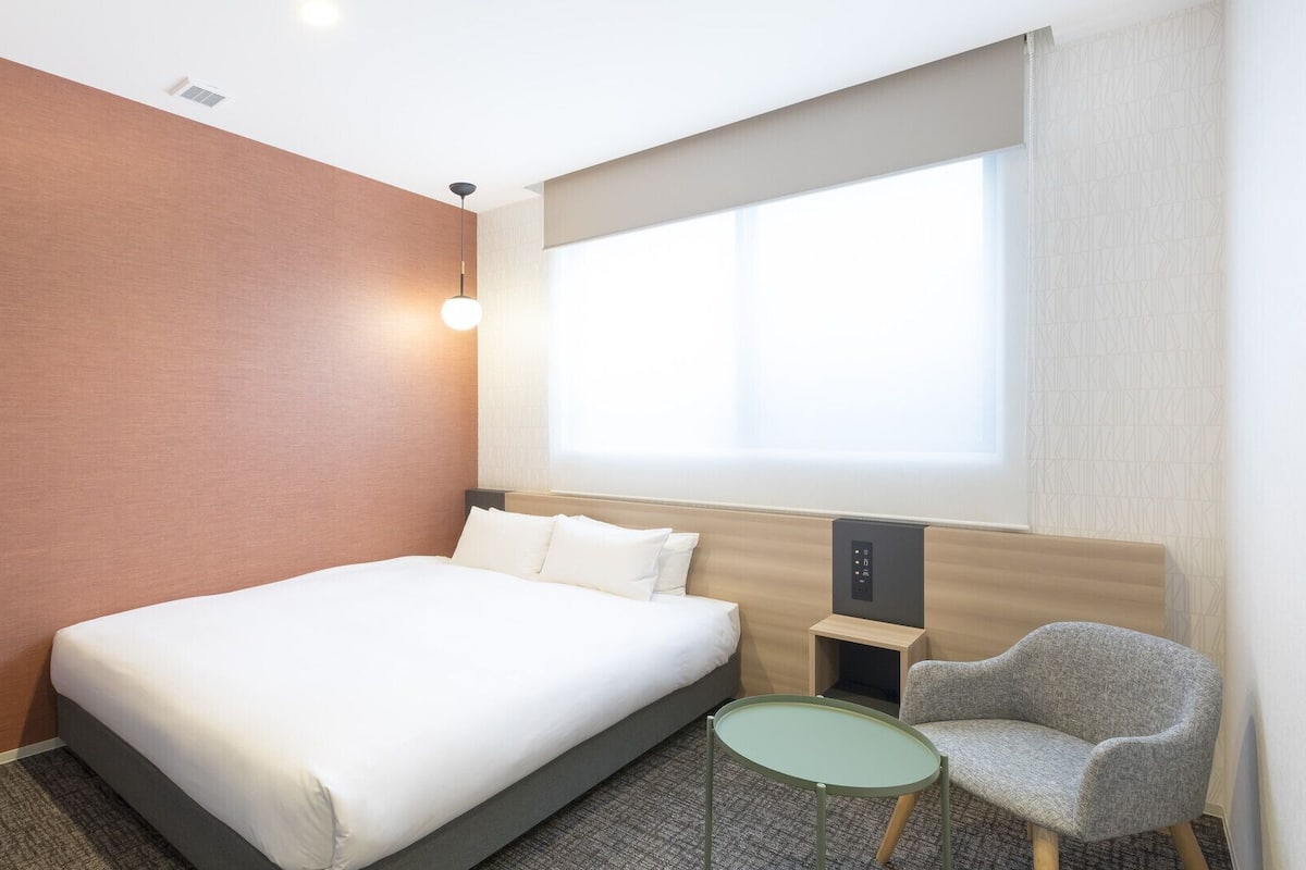 A hotel room with a bed, a small table, a chair, and a window with curtains.