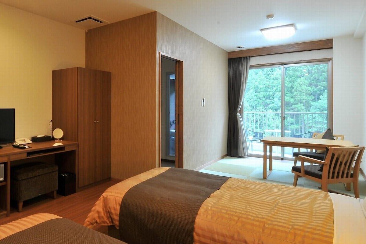 A hotel room with two beds, a desk with a computer, a chair, and a balcony with a view of trees.