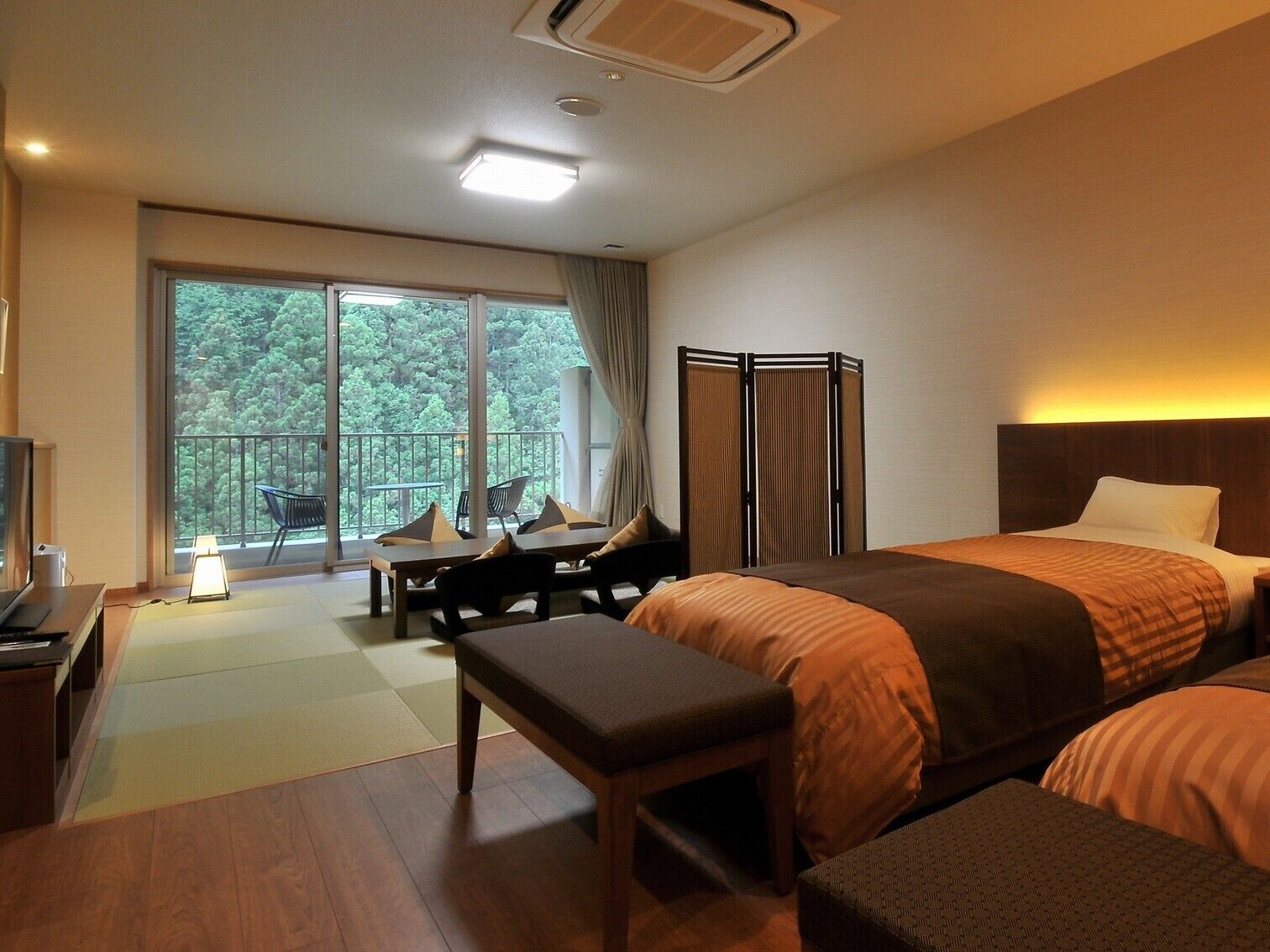 A hotel room with a bed, a desk, a chair, a bench, a TV, and a balcony with a view of trees.