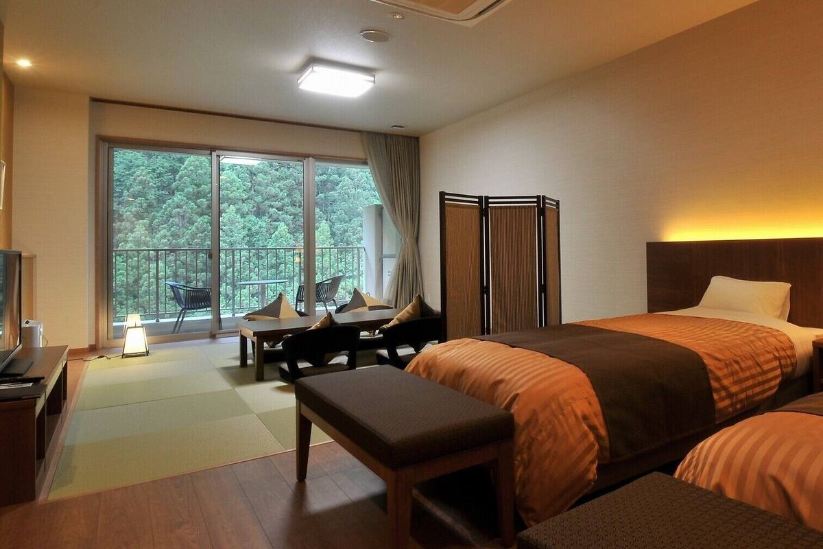 A hotel room with a bed, a desk, a chair, a bench, a TV, and a balcony with a view of trees.