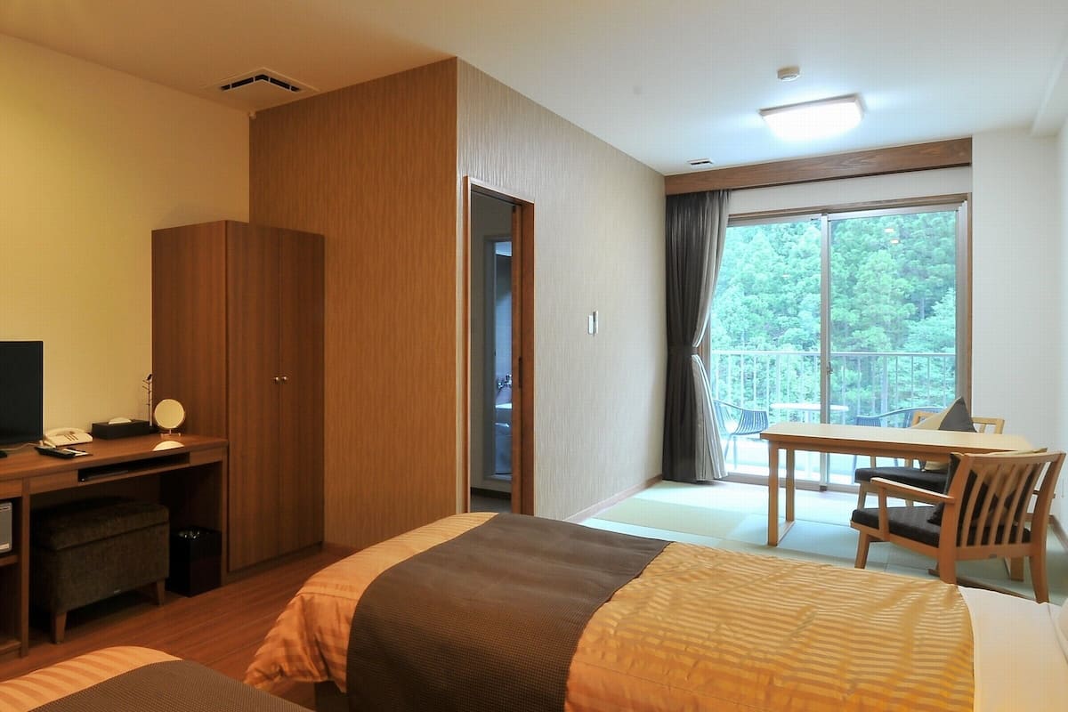 A hotel room with two beds, a desk, a chair, a TV, and a balcony with a view of trees.