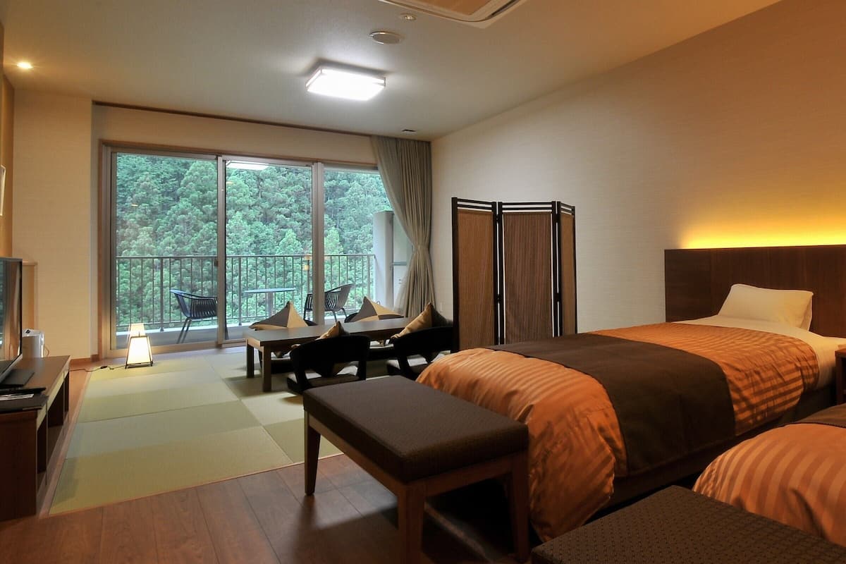 A hotel room with a bed, a desk, a chair, a bench, a TV, and a large window with a view of trees.