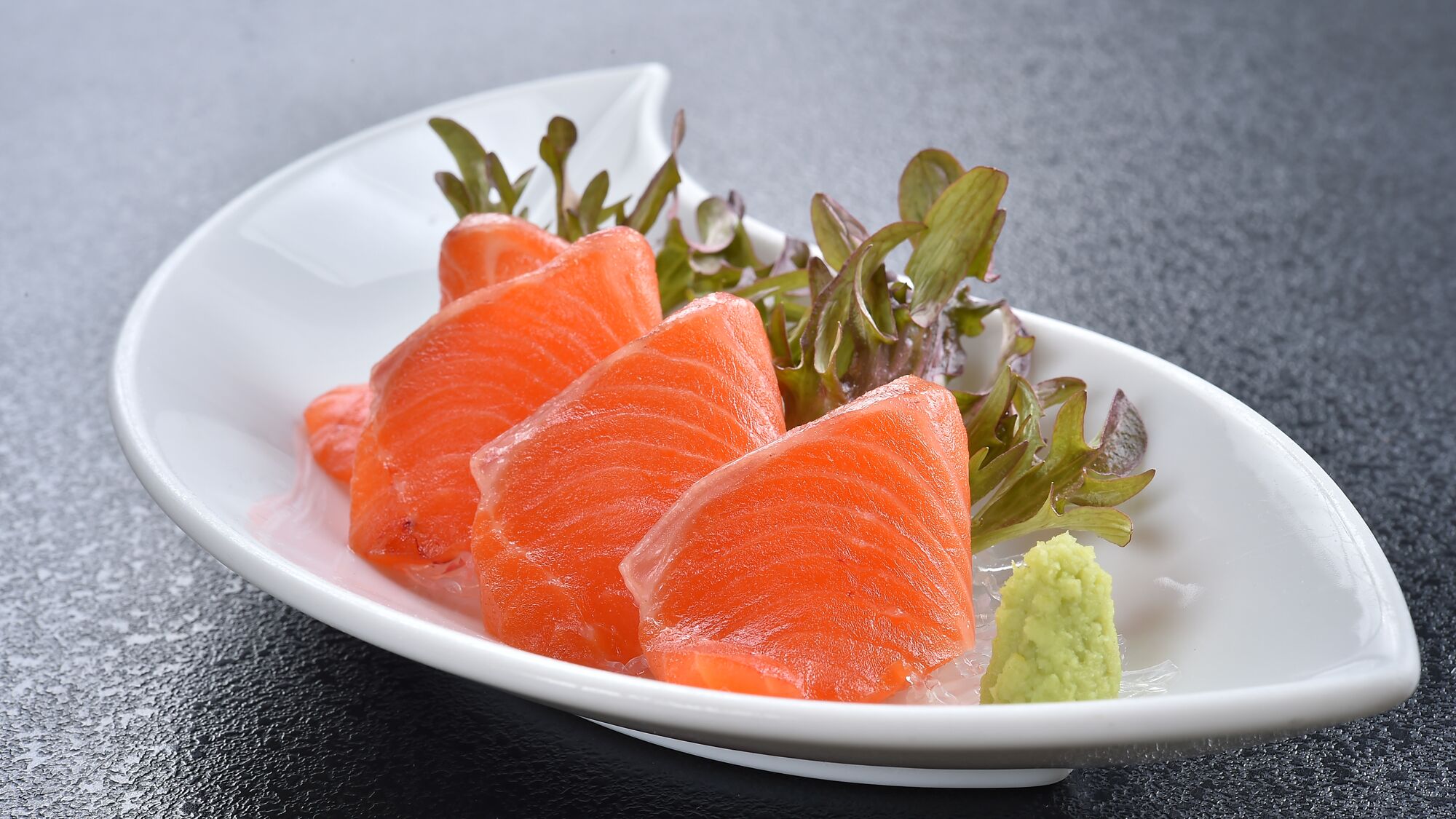 Une assiette blanche sur laquelle on voit des tranches de saumon, une cuillerée de sauce verte et quelques feuilles de verdure, le tout posé sur une surface sombre.