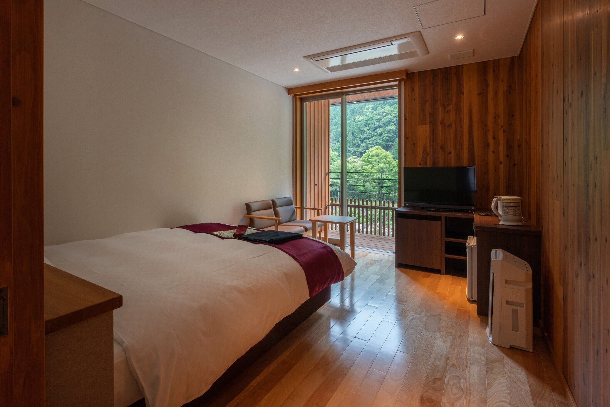 Une chambre avec un lit, un bureau, une télévision et un balcon donnant sur les arbres.