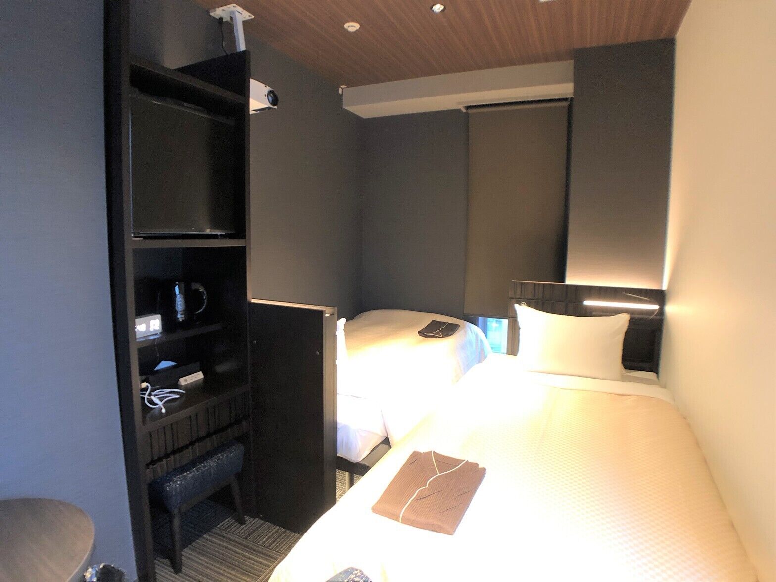 A hotel room with two beds, a desk, a bookshelf, and a ceiling-mounted projector.