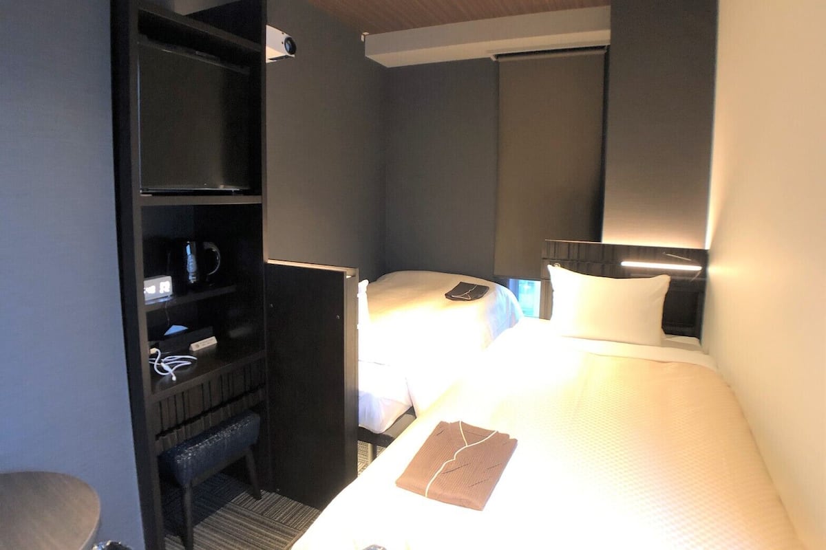 A hotel room with two beds, a desk, a bookshelf, and a ceiling-mounted projector.
