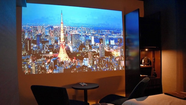 A hotel room with a bed, a small table, and a wall-mounted screen displaying a cityscape. - J Hotel Tokyo Geo (Tokyo)