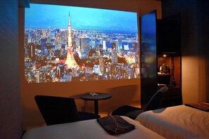 A hotel room with a bed, a small table, and a wall-mounted screen displaying a cityscape. - J Hotel Tokyo Geo (Tokyo)