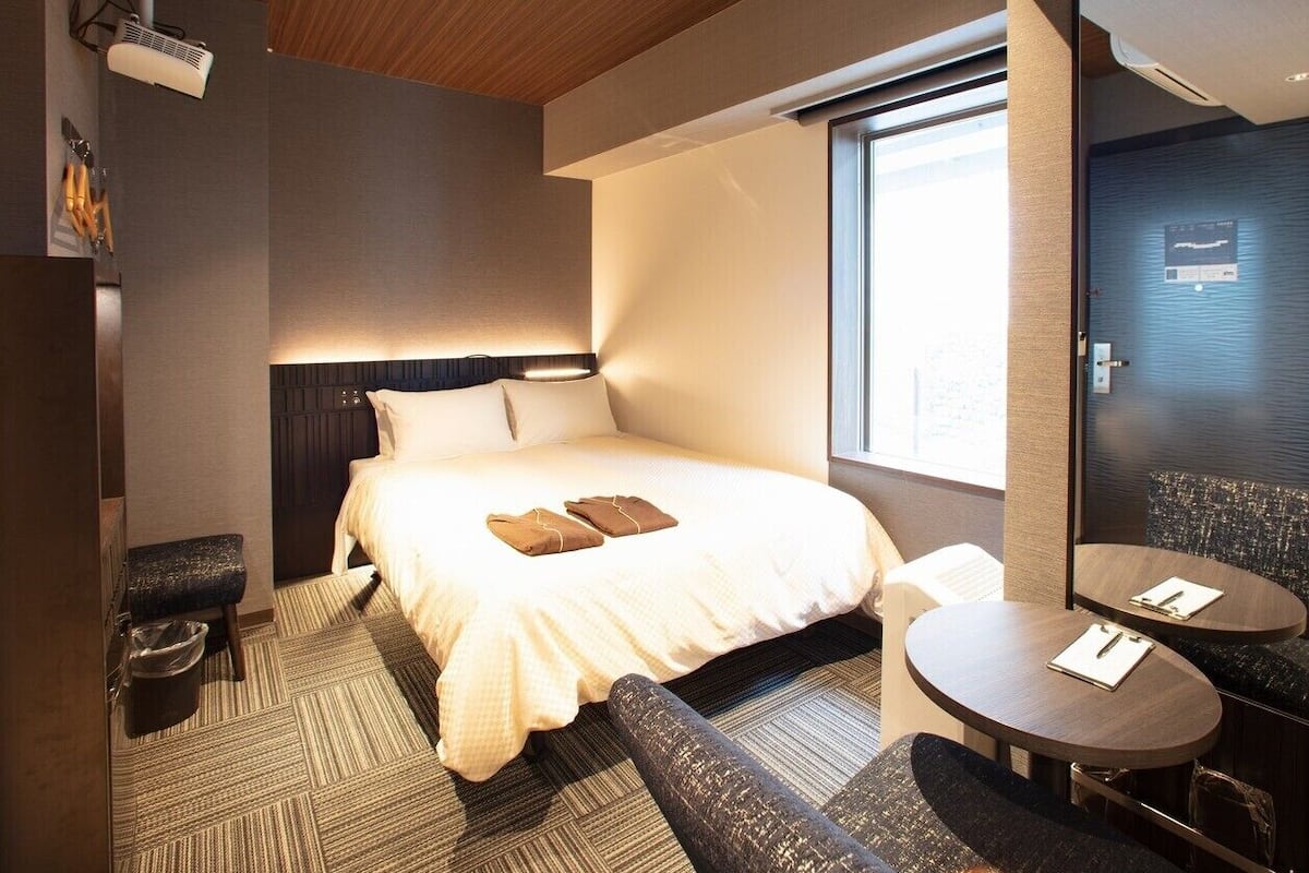 A hotel room with a bed, bedside table, chair, and a large window.