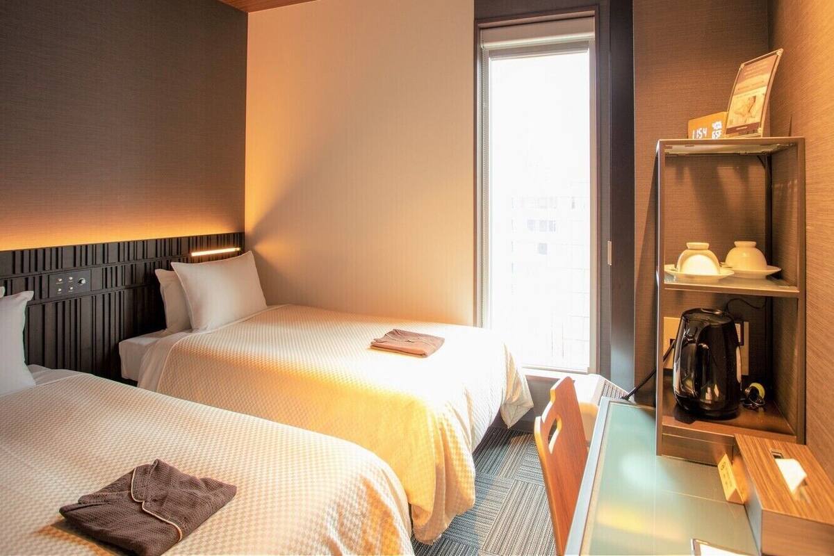 A hotel room with two beds, a desk, a coffee machine, and a shelf with a picture frame and a cup.