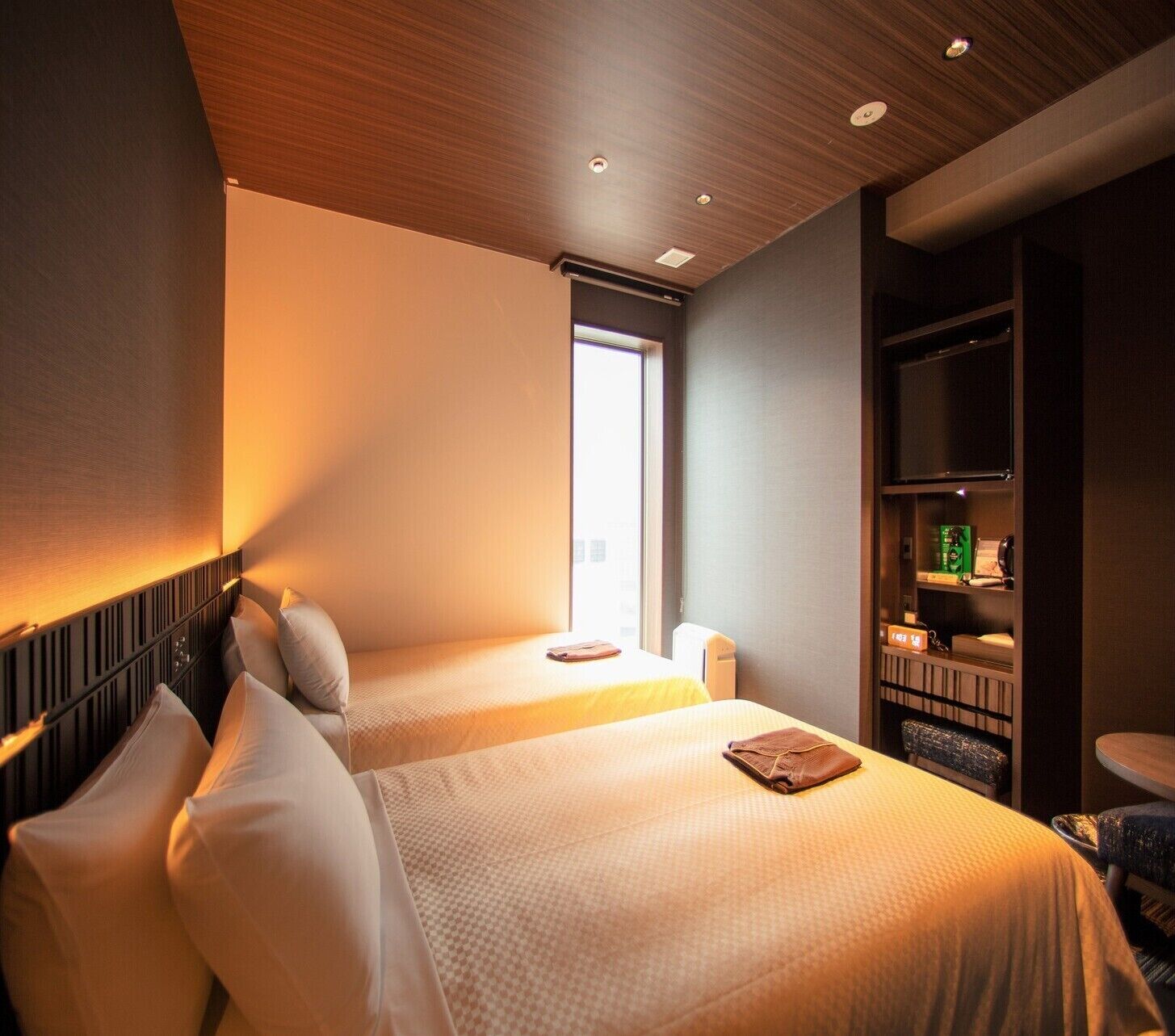 A hotel room with two beds, a window, a bookshelf, and a small table.
