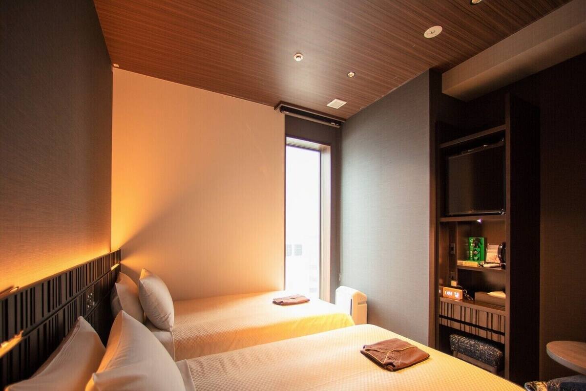 A hotel room with two beds, a window, a bookshelf, and a small table.