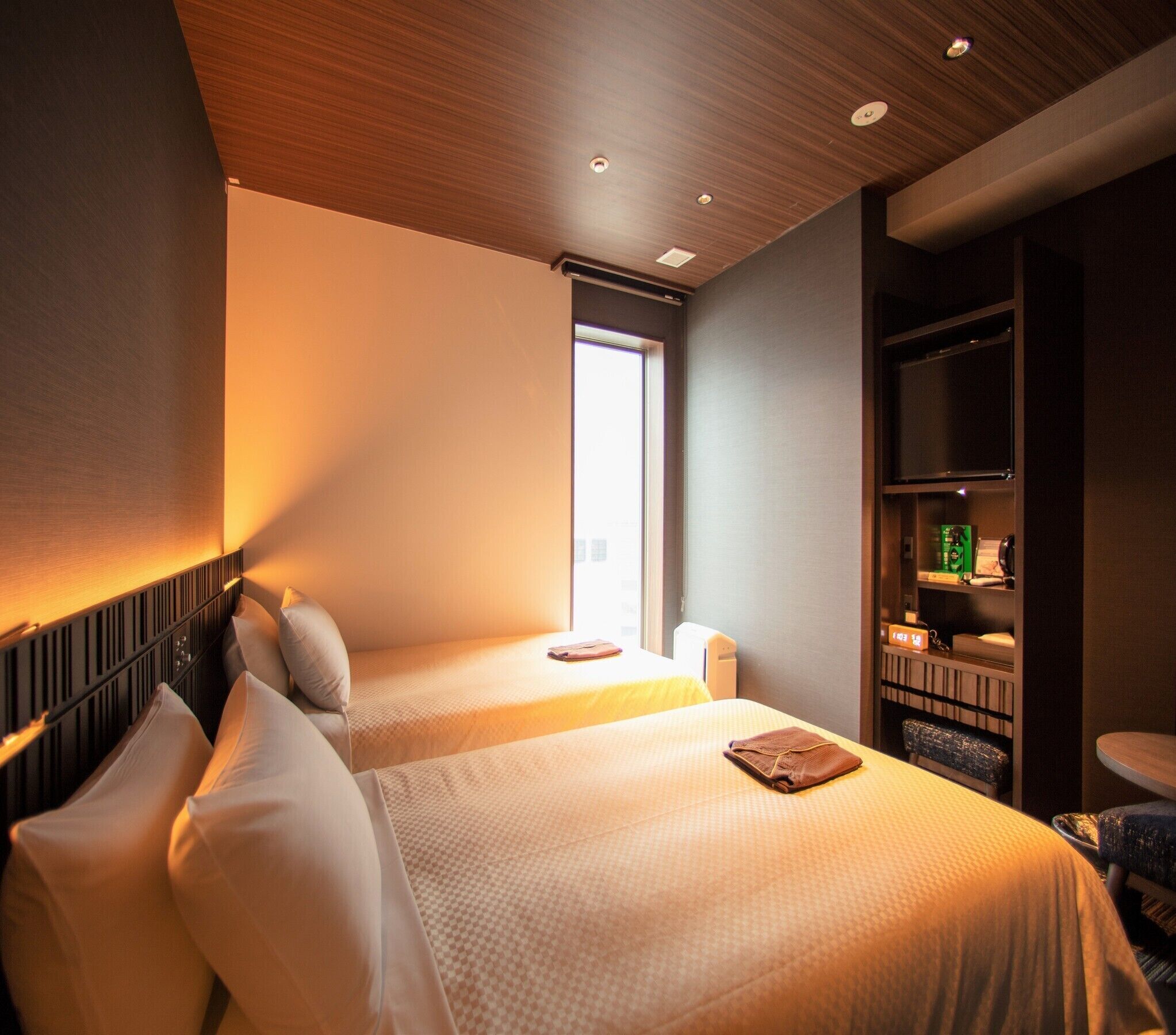 A hotel room with two beds, a built-in shelf with toiletries, and a window providing natural light.