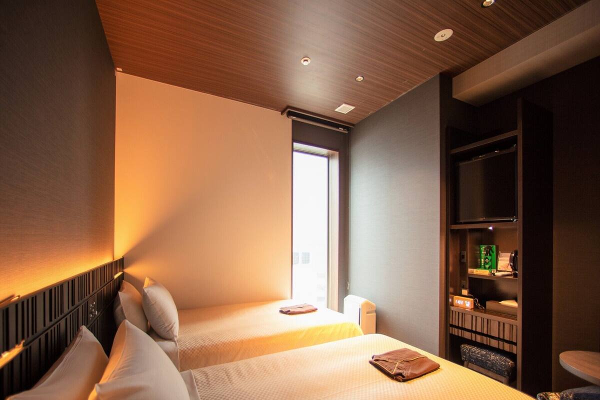 A hotel room with two beds, a built-in shelf with toiletries, and a window providing natural light.