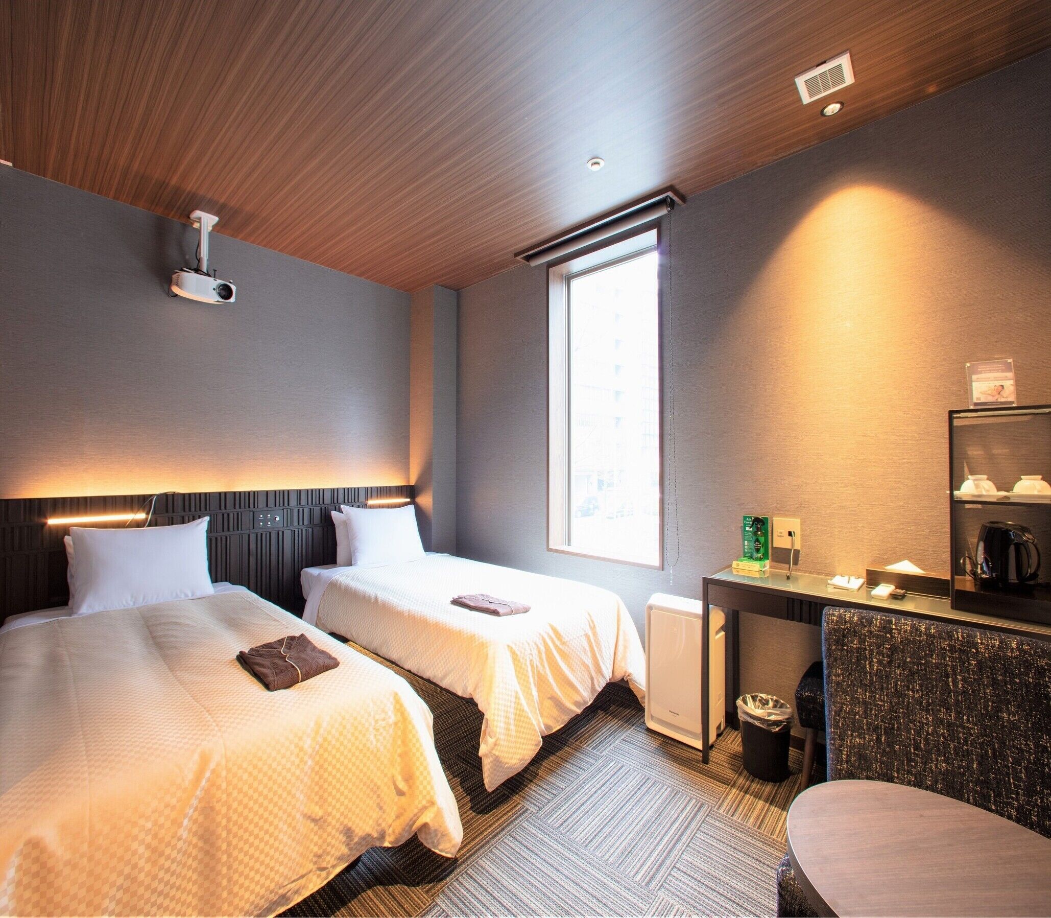 A hotel room with two beds, a desk, a chair, a radiator, a projector, a window, and a trash bin.