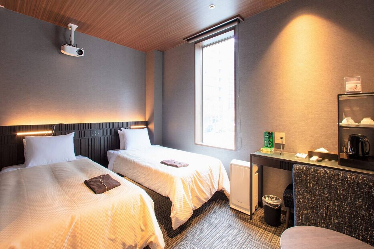 A hotel room with two beds, a desk, a chair, a radiator, a projector, a window, and a trash bin.