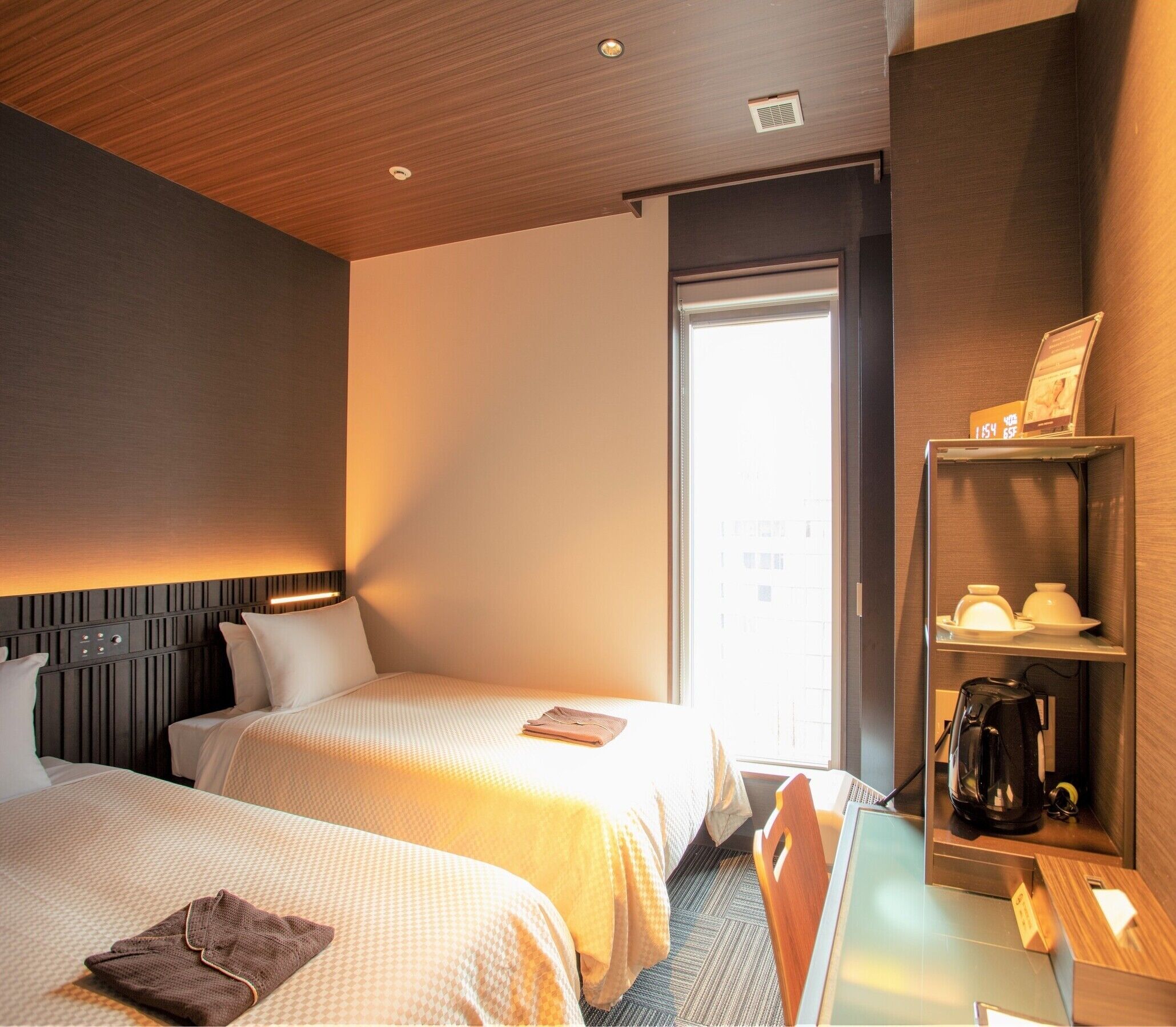 A hotel room with two beds, a desk, a coffee maker, and a shelf with a kettle.