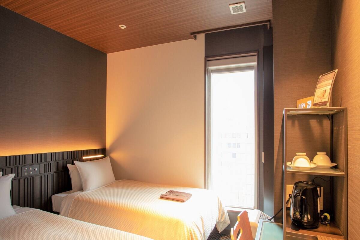 A hotel room with two beds, a desk, a coffee maker, and a shelf with a kettle.