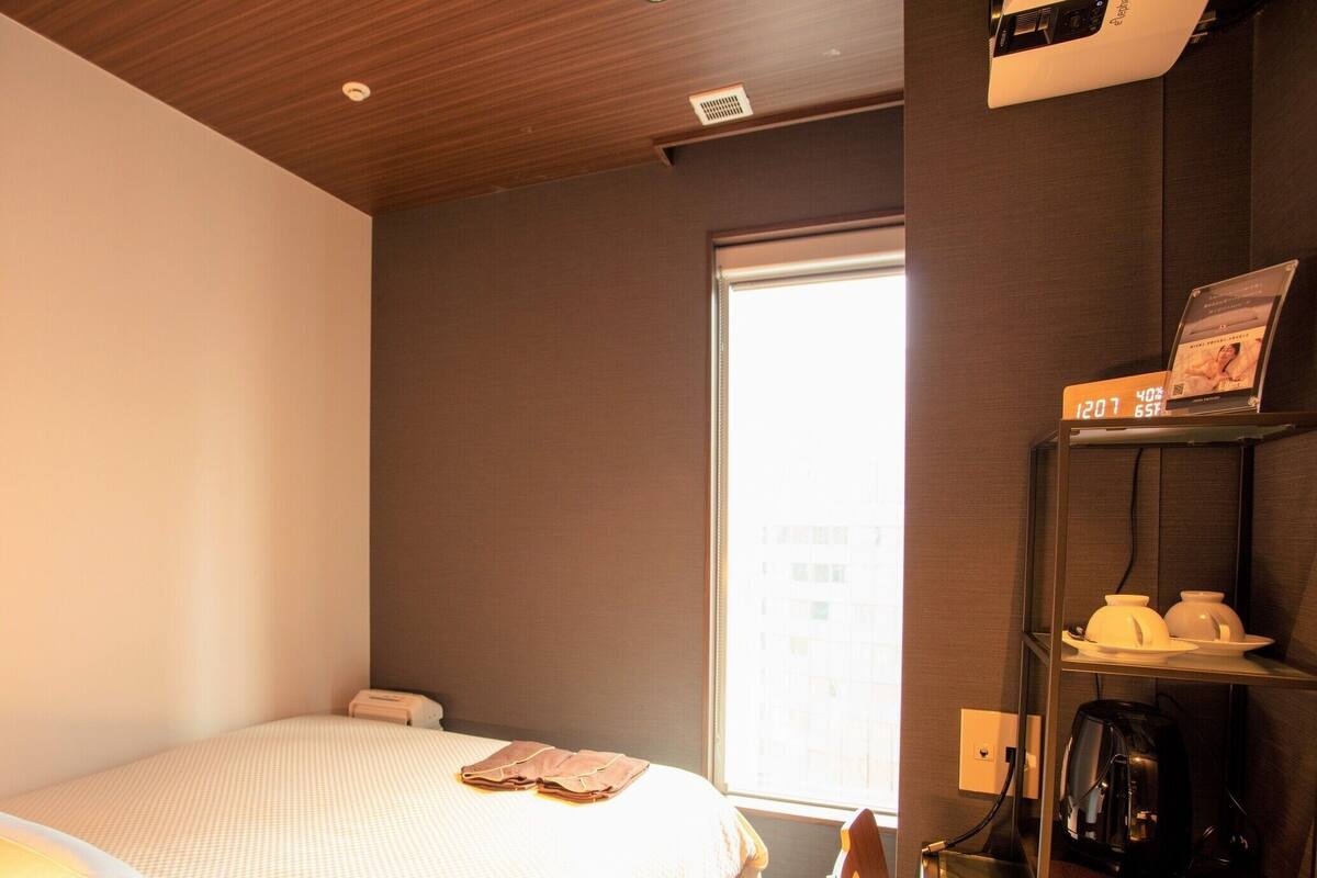 A hotel room with a bed, a desk with a coffee machine, a shelf with toiletries, and a window with a view of buildings.