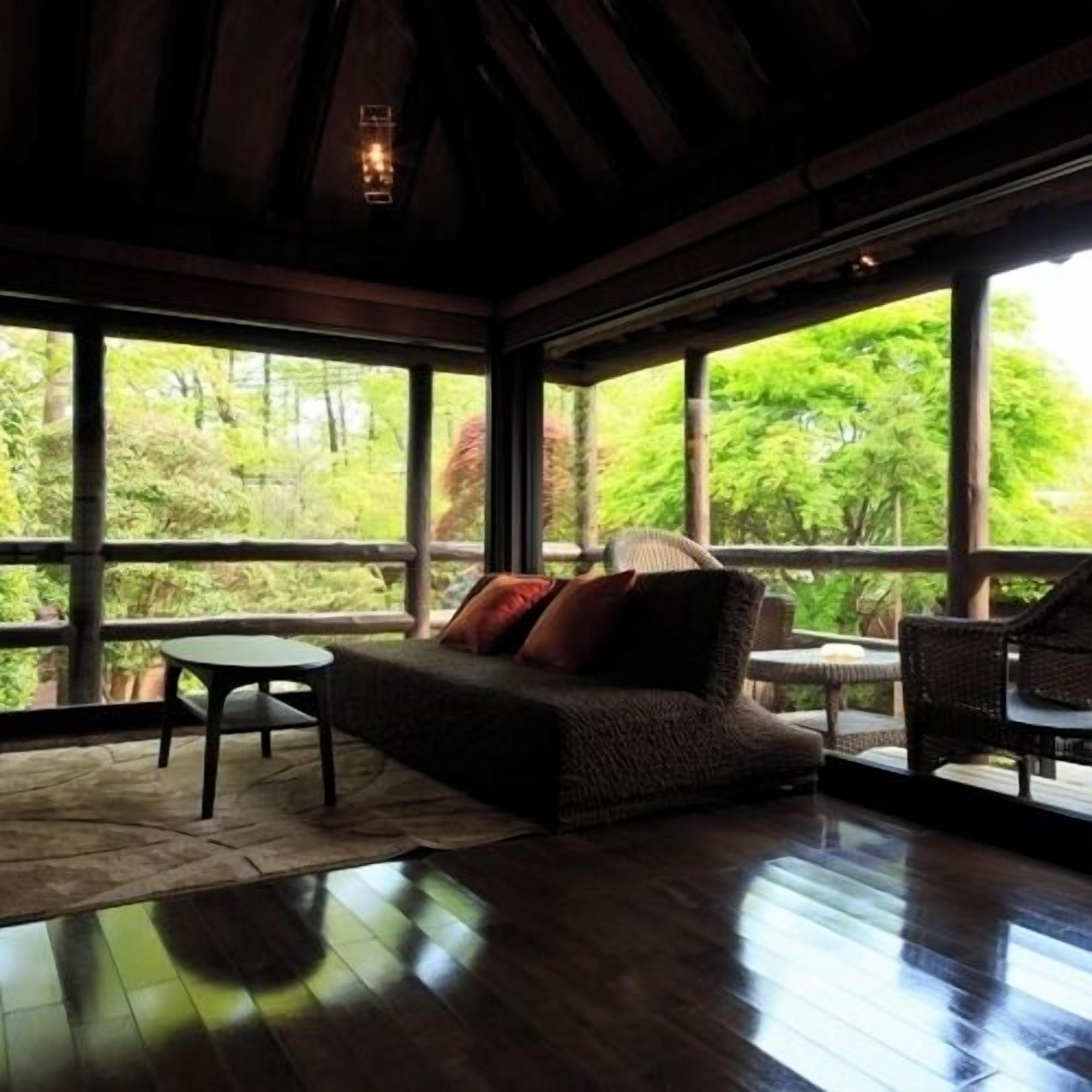 Una habitación con piso de madera, un sofá con cojines, una mesa y sillas. Amplias ventanas ofrecen vistas al verdor exterior.
