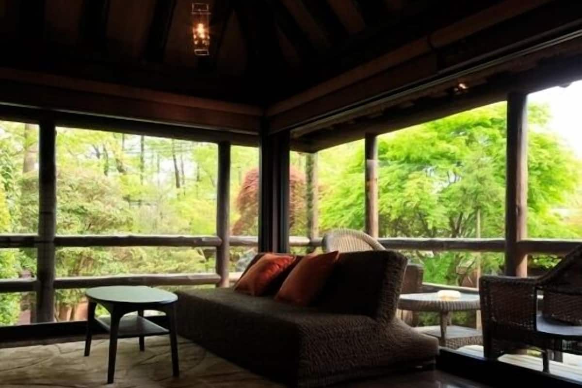 Una habitación con sofá, una mesa y sillas, ventanas amplias con vistas a zonas verdes.