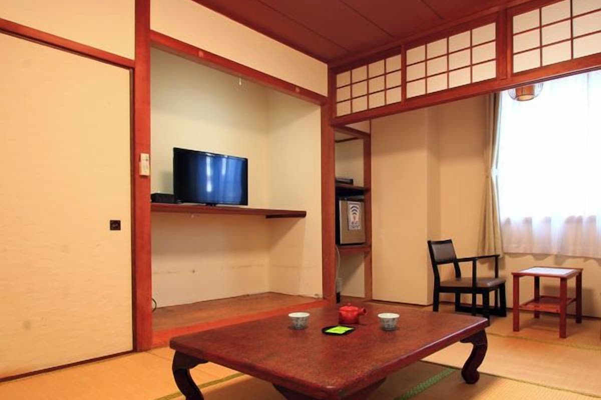 A traditional Japanese room with tatami flooring, a low wooden table, a television on a shelf, and a sliding door.