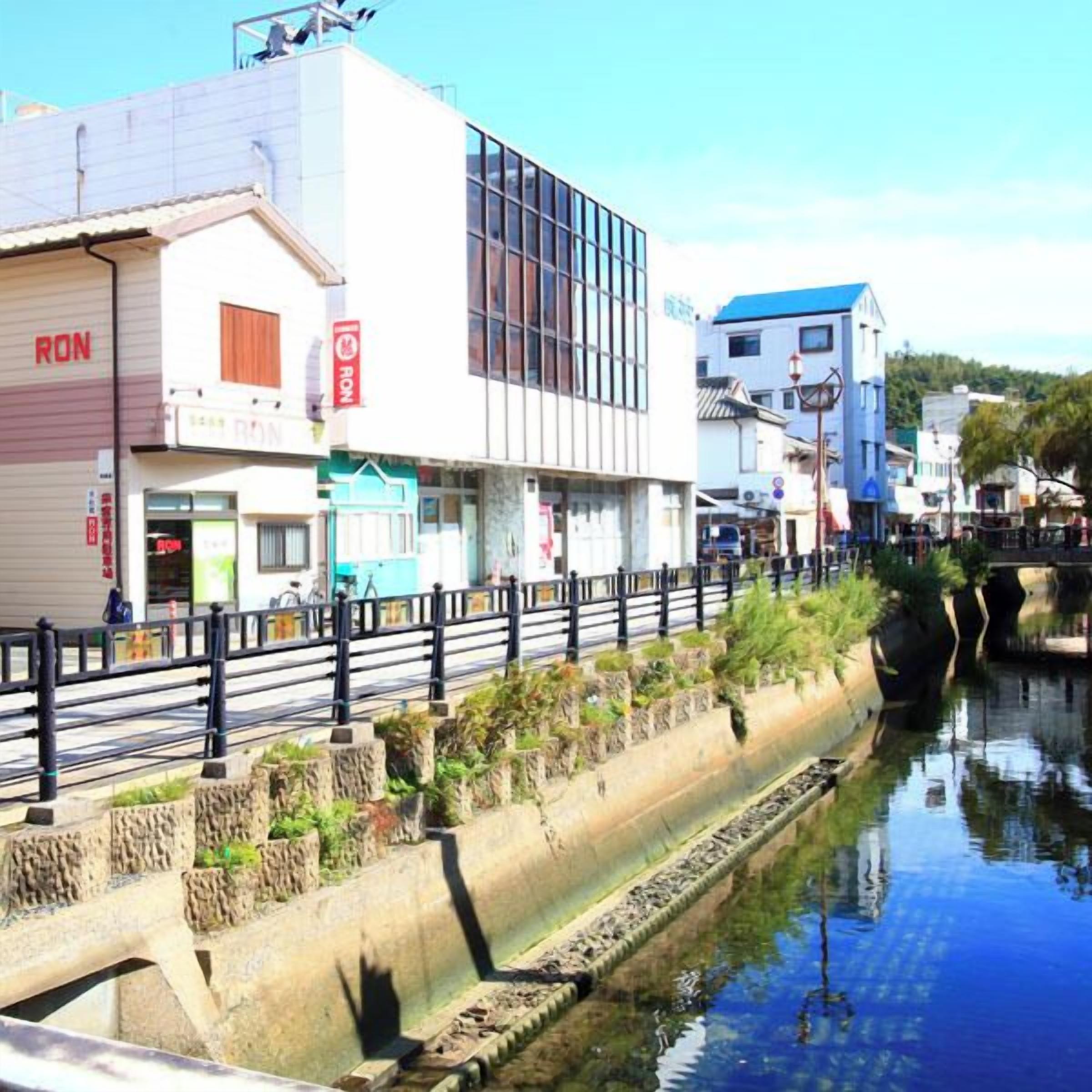 A riverside walkway with a concrete barrier, a bridge, and buildings with signage including "RON".