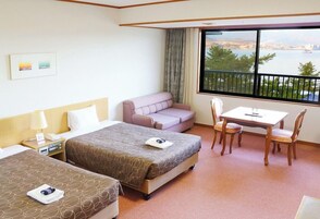 Habitación de hotel con dos camas, un sofá, una mesa con sillas, una ventana que ofrece vistas al agua y al paisaje urbano, y una mesita de noche con lámpara y teléfono.