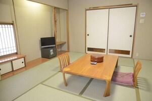 Una habitación japonesa tradicional con suelo de tatami, una mesa de madera, sillas, un televisor y puertas correderas.