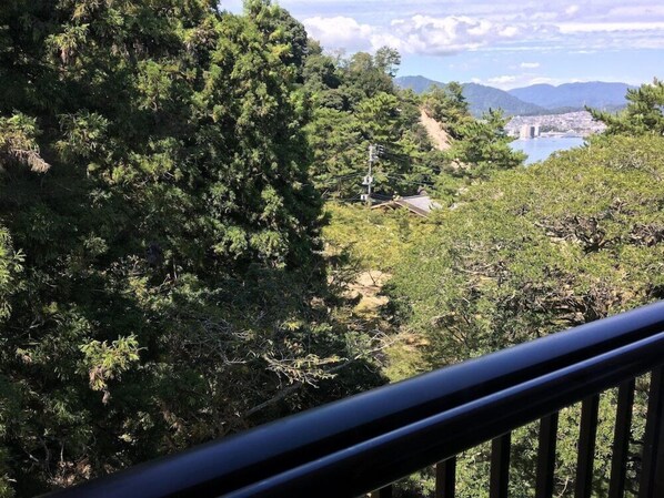 A view from a balcony with dense foliage, a distant town, and a body of water. - Miyajima Morinoyado (Hatsukaichi)