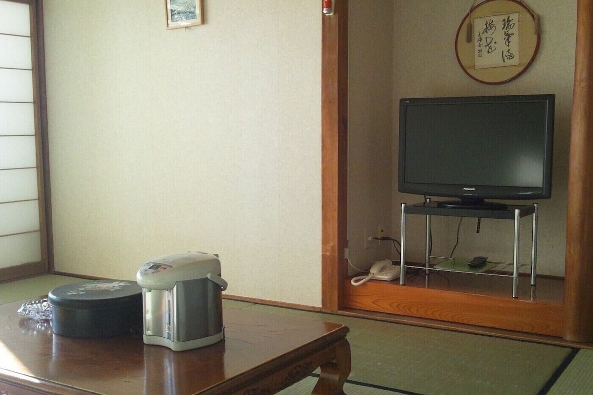 A room with a wooden table, a coffee maker, a television on a stand, and a traditional Japanese tatami mat floor.