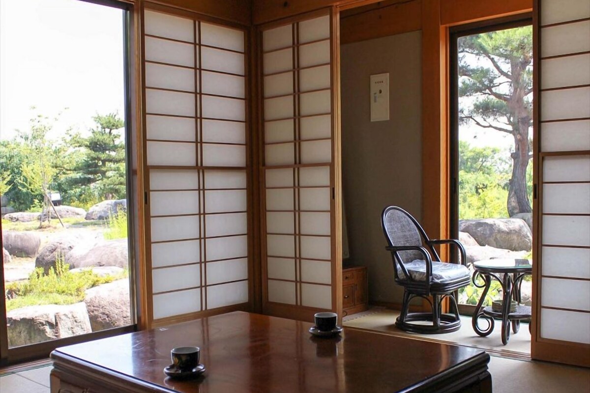 Une pièce de style japonais traditionnel, avec un sol recouvert de tatamis, une petite table en bois, une chaise et une vue sur un jardin à travers des panneaux coulissants en papier.