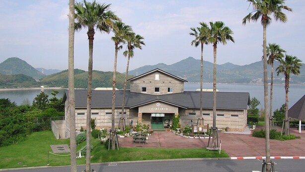 A building with a stone facade, a central entrance, and a view of a body of water and mountains in the background. - Green Stay Nagaura (Suooshima)