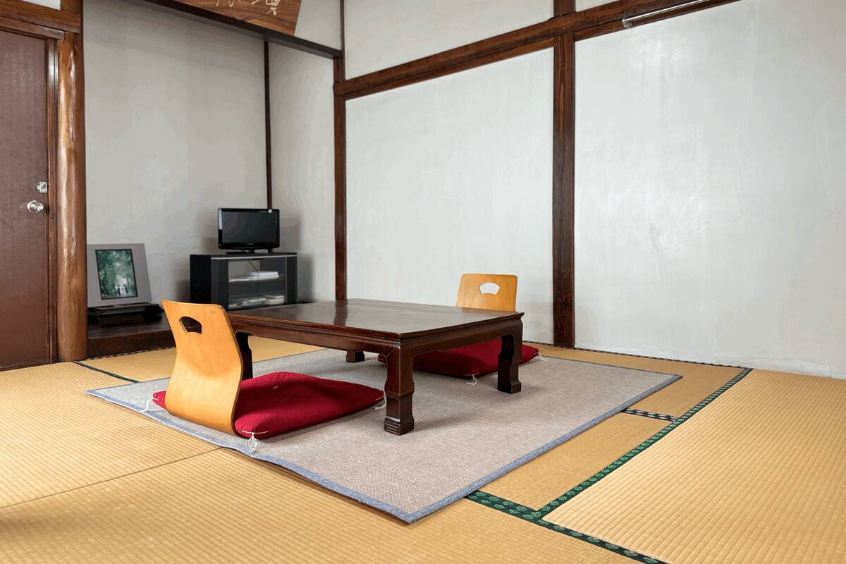 A traditional Japanese room with tatami flooring, a low wooden table, two wooden chairs, a television, and a cabinet.