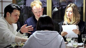 Un grupo de personas sentadas alrededor de una mesa, con copas de vino blanco, conversando.