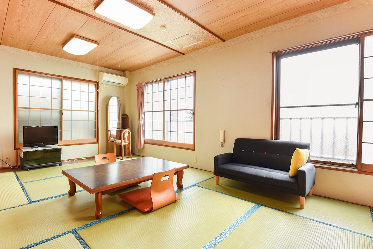 A room with tatami flooring, a sofa, a table, a TV, and large windows.