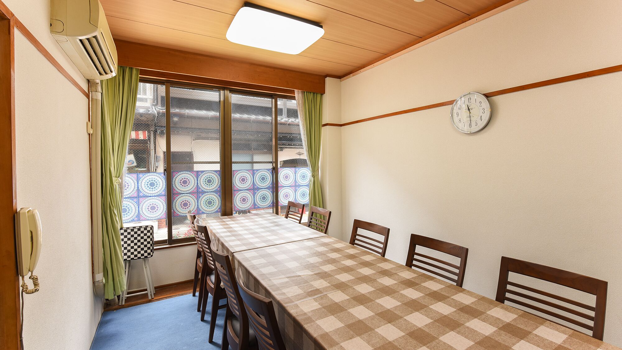 A room with a checkered table, wooden chairs, a clock on the wall, and a window with patterned curtains.