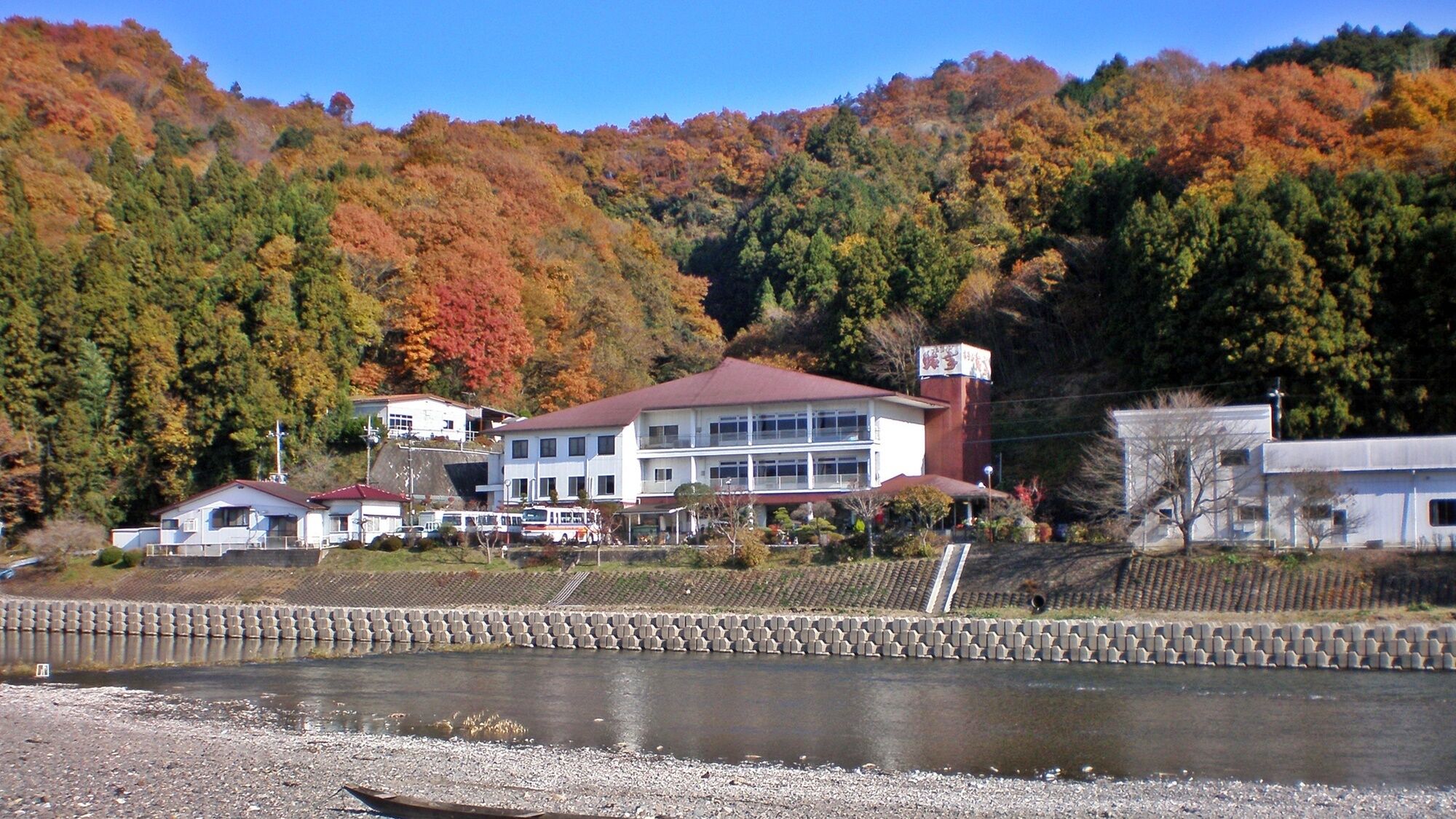 Un edificio lungo un fiume con tetto rosso, circondato da alberi dai colori autunnali.