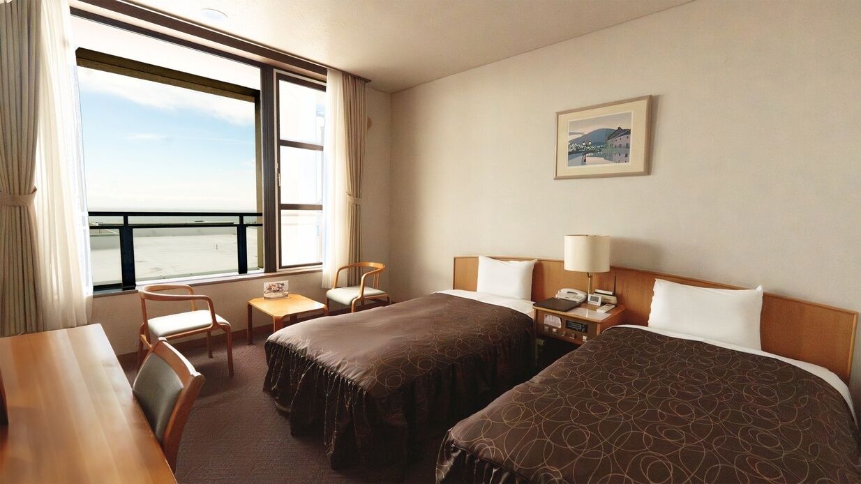 Habitación de hotel con dos camas, un escritorio, una silla, un ventanal con vistas y un cuadro en la pared.