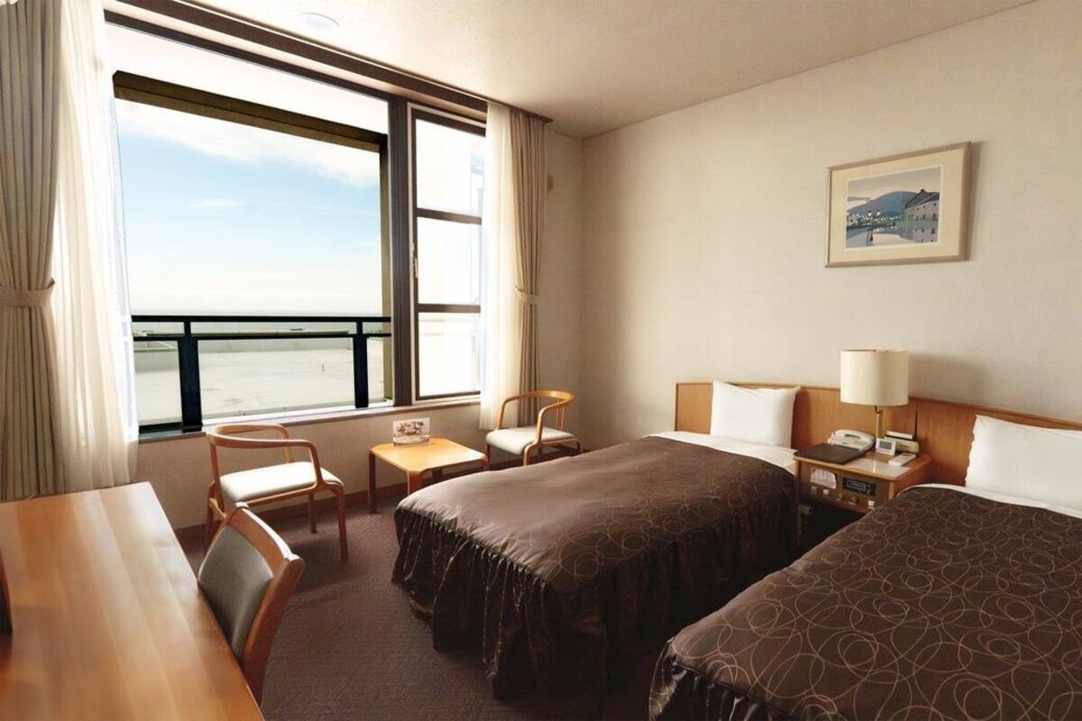 Habitación de hotel con dos camas, un escritorio, una silla, un ventanal con vistas y un cuadro en la pared.