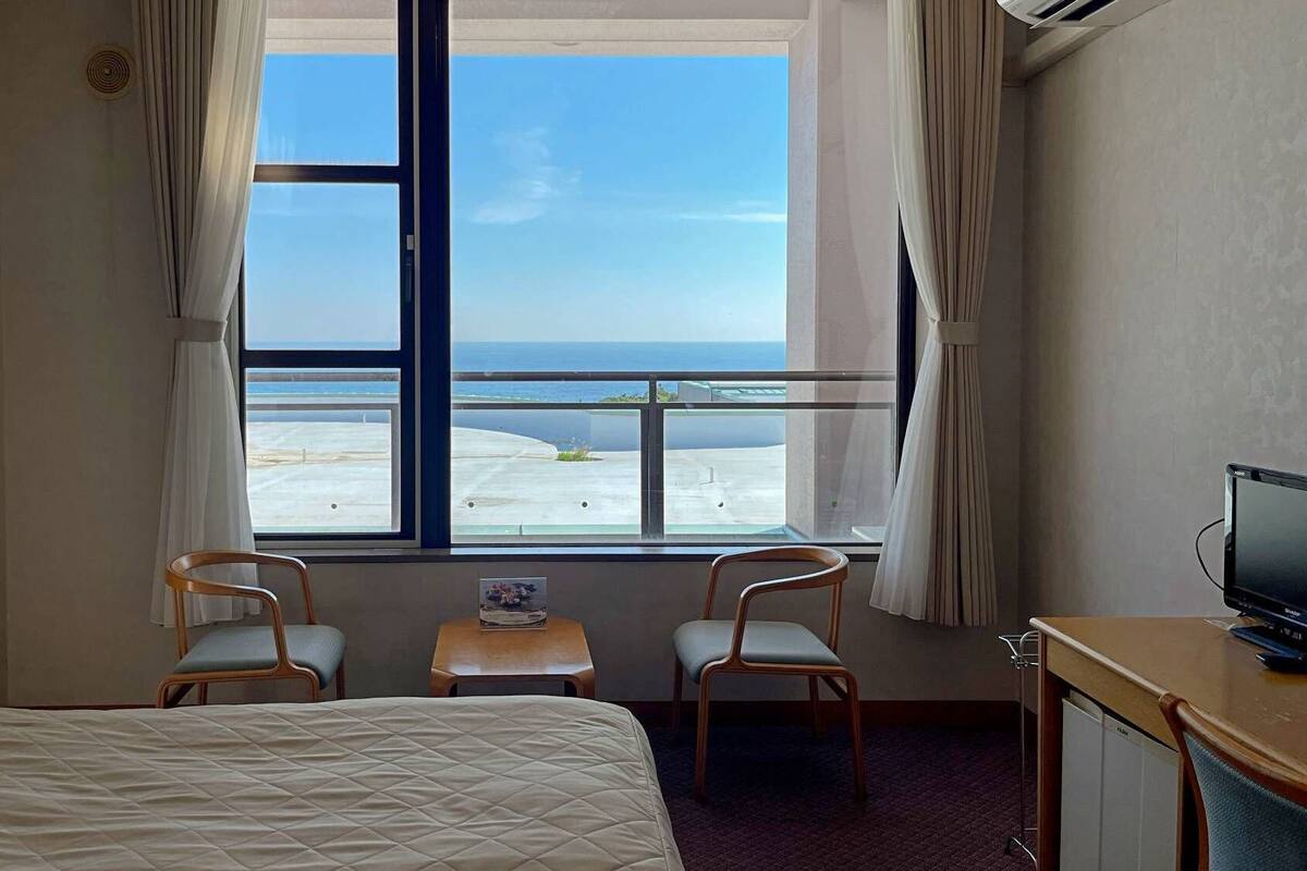 Habitación de hotel con cama, dos sillas, una mesita, televisión y ventana con vista a la playa.
