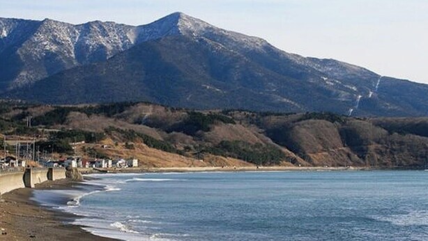 Paisaje costero con una montaña al fondo, una playa y una estructura con techo curvo.