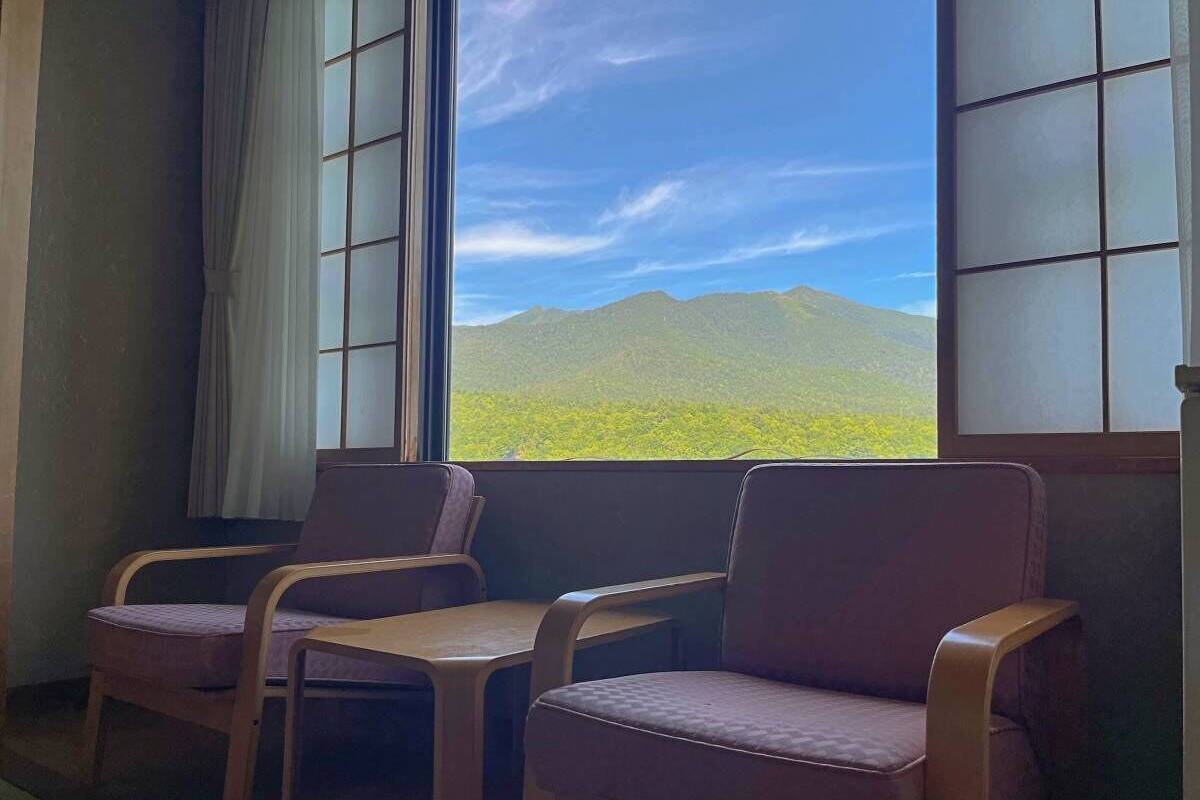 Una habitación con vistas a verdes colinas a través de una ventana, tres sillas de madera y una mesa.