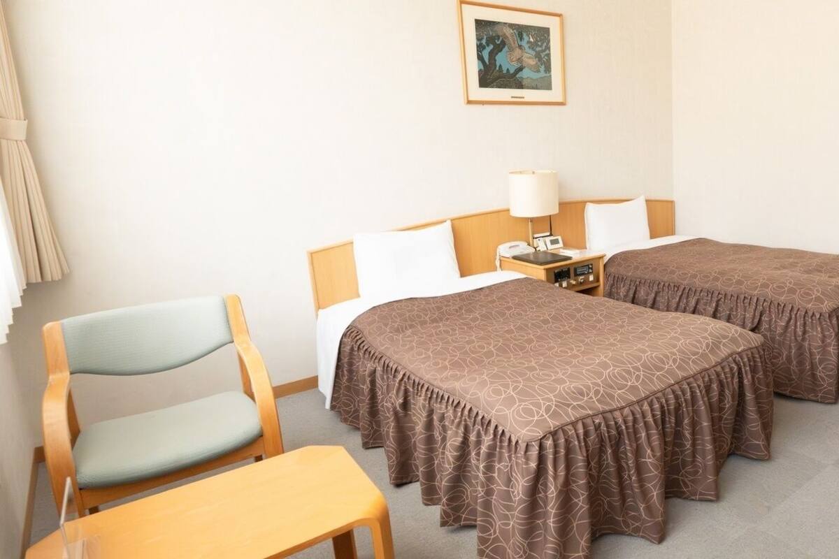 Habitación de hotel con dos camas, una silla, una mesa, una lámpara y un cuadro en la pared.