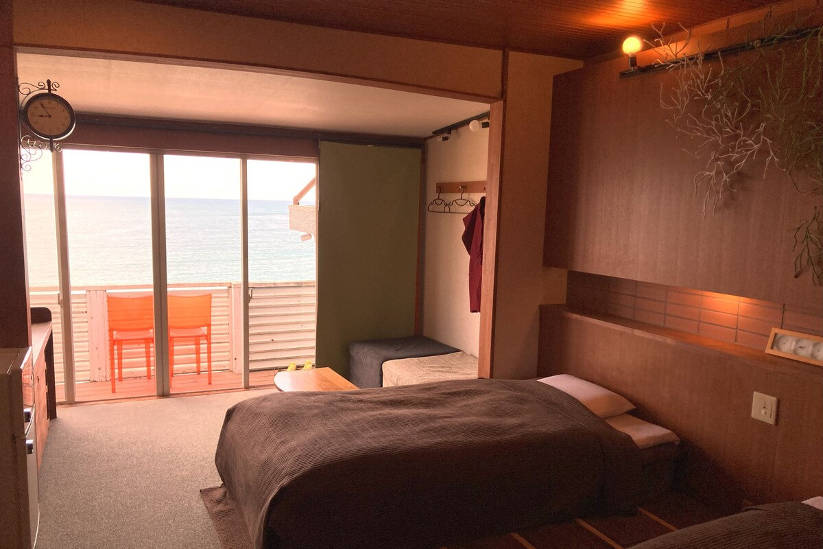A bedroom with a bed, a small table, and a chair. There is a view of the sea through a sliding door.